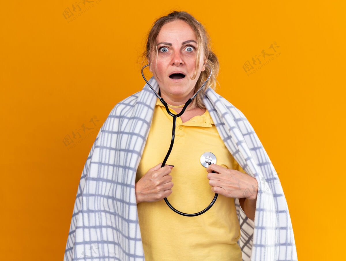 担心不健康的女人裹着毯子 感觉不舒服 患流感和感冒 站在橘色的墙上 用听诊器听心跳 看起来很担心毯子心跳流感