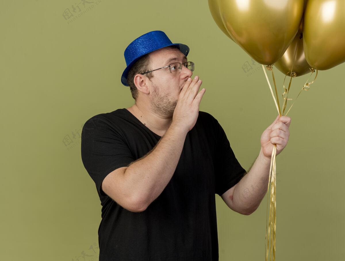 橄榄自信的成年斯拉夫人戴着眼镜 戴着蓝色派对帽 手贴着嘴看着氦气球穿眼镜帽子