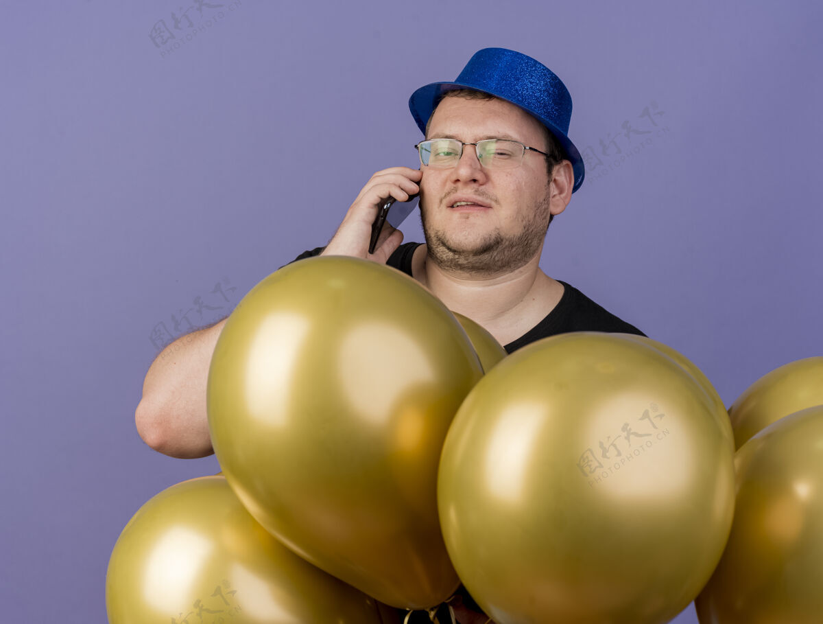 电话自信的成年斯拉夫人戴着眼镜 戴着蓝色派对帽 手持氦气球站在那里打电话光学氦成人