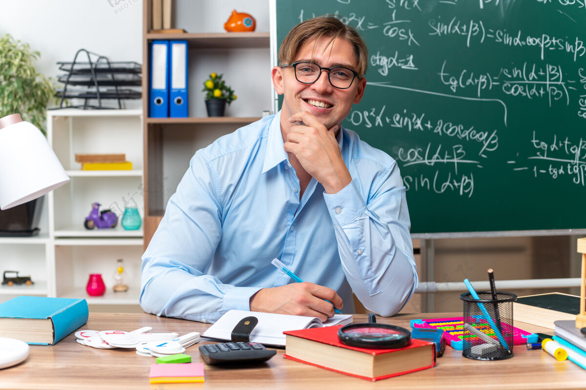 笔记快乐微笑的年轻男老师坐在课桌旁 在教室的黑板前拿着书 笔和笔记年轻男人书桌