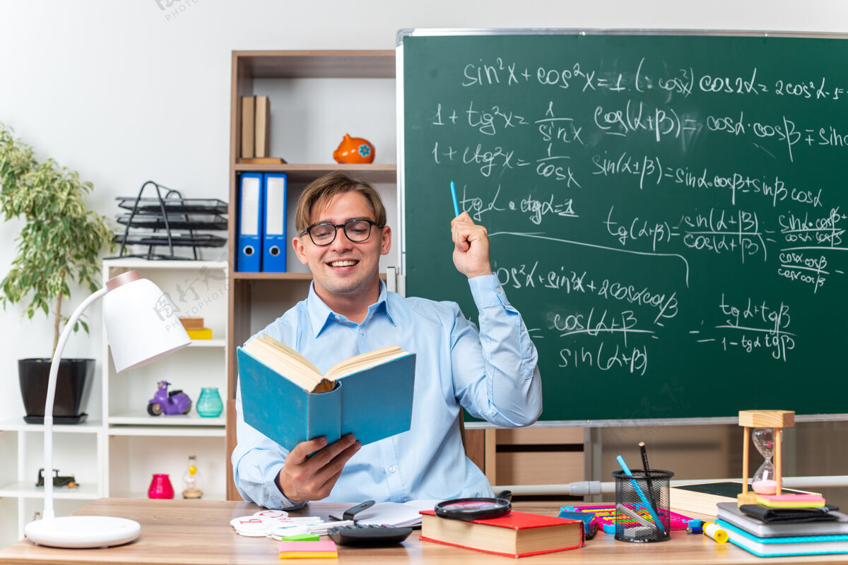 阅读年轻的男老师戴着眼镜坐在书桌旁 拿着书和笔记 在教室的黑板前愉快地 积极地笑着看书快乐欢呼男性