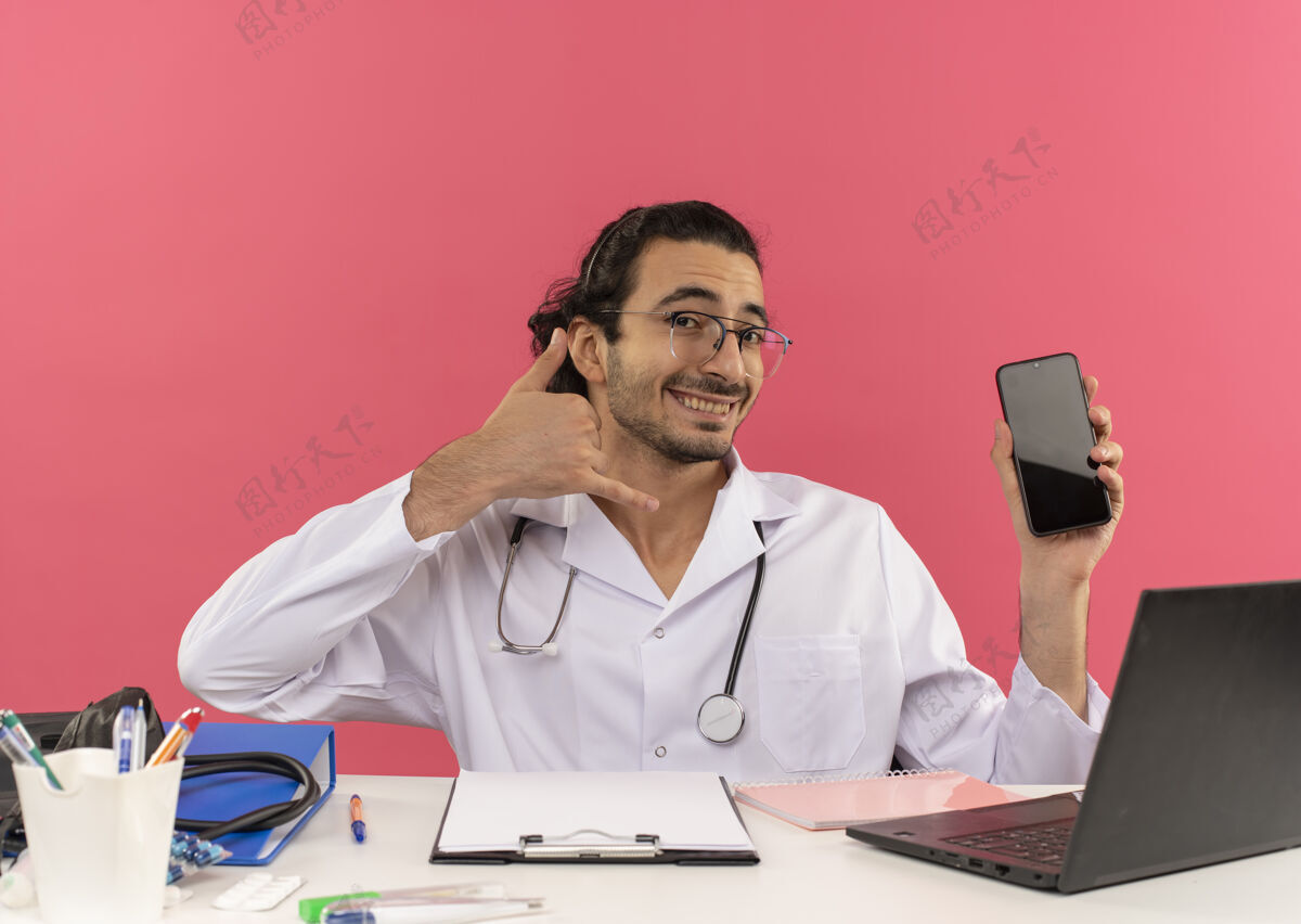年轻人面带微笑的年轻男医生 戴着医用眼镜 穿着医用长袍 听诊器坐在办公桌旁空间展示桌子