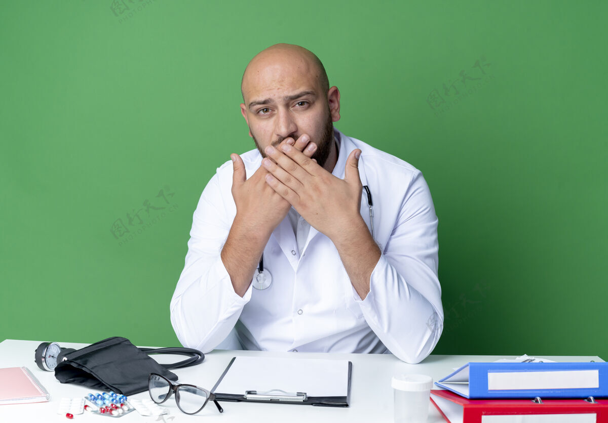 嘴年轻的男医生穿着医用长袍和听诊器坐在办公桌旁坐着工具穿