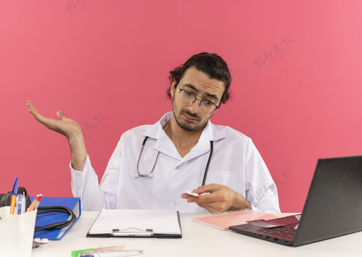 粉红色困惑的年轻男医生戴着医用眼镜 穿着医用长袍 听诊器坐在办公桌旁点眼镜长袍