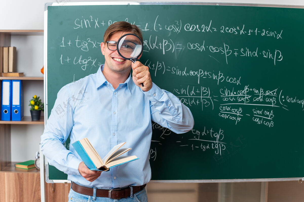 玻璃年轻的男老师戴着眼镜 拿着放大镜和书 兴高采烈地在教室里用数学公式站在黑板旁边讲解功课年轻快乐公式
