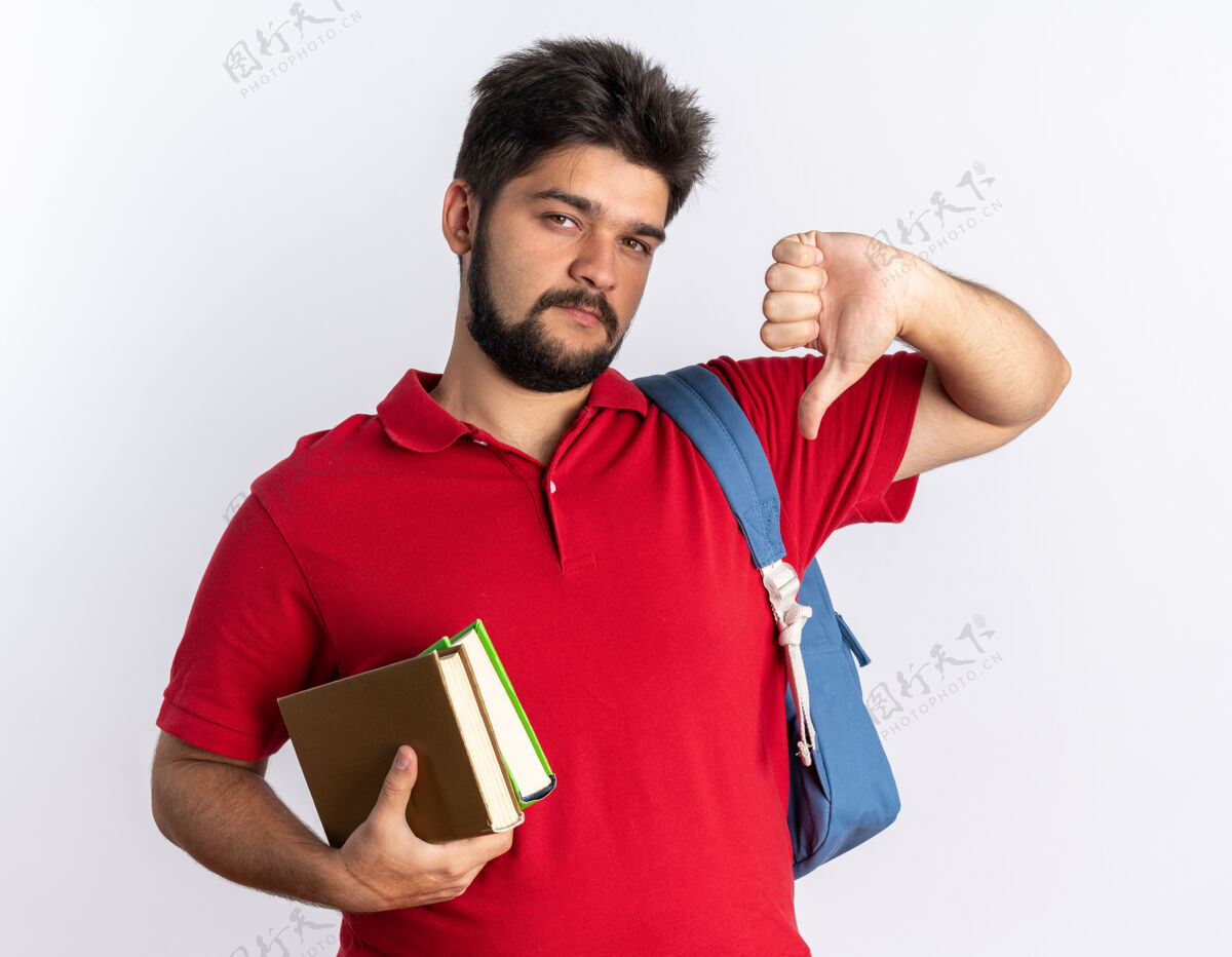 胡子年轻的留着胡子的学生 穿着红色马球衫 背着背包 拿着笔记本 看起来很不高兴 站着竖起大拇指站着笔记本不高兴