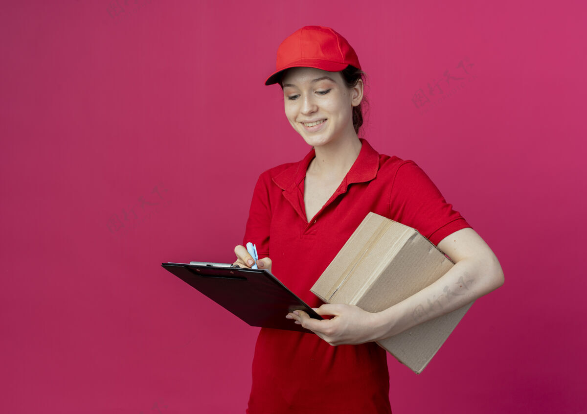 年轻微笑着的年轻漂亮的送货女孩 穿着红色制服 戴着帽子 拿着剪贴板 用笔在剪贴板上写字微笑钢笔交付
