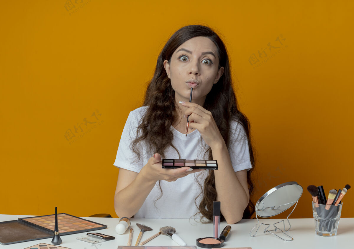 女孩印象深刻的年轻漂亮的女孩坐在化妆台上 化妆工具拿着眼影调色板 用眼影笔抚摸嘴唇坐刷子年轻