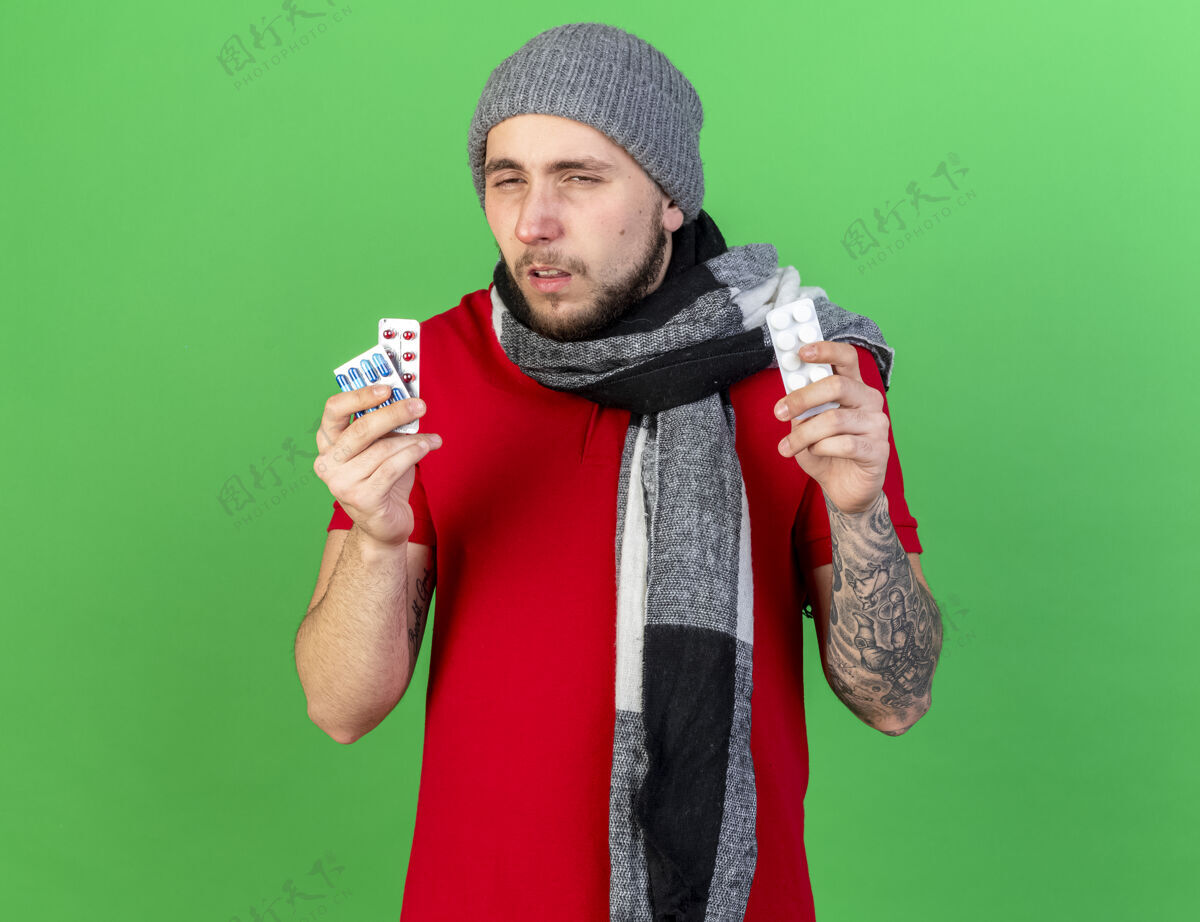 穿苍白的年轻白种人 戴着冬天的帽子 戴着围巾 手里拿着绿色的药片年轻人药片男人