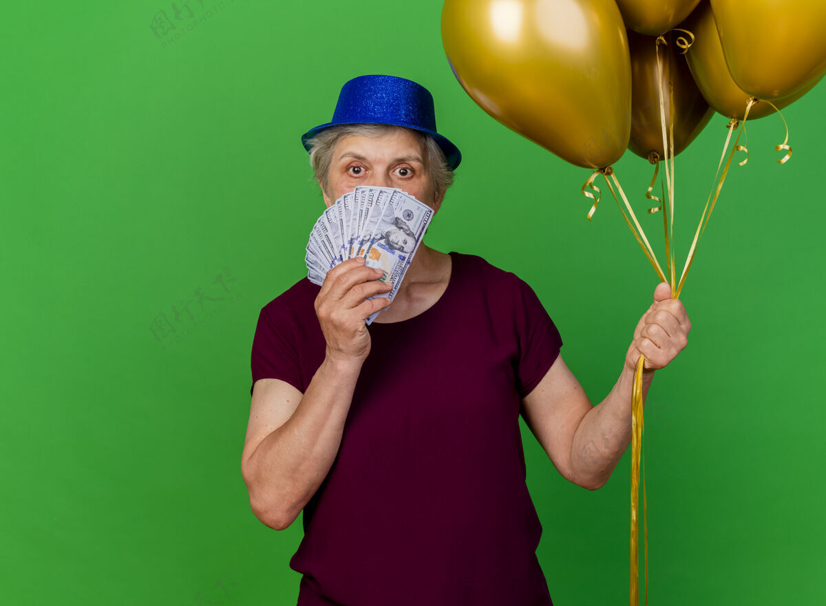 老人惊讶的老妇人戴着聚会的帽子 手里拿着钱和氦气球在果岭上帽子氦惊喜