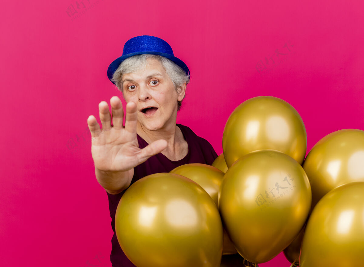 粉色惊讶的老妇人戴着派对帽站着 手里拿着氦气球 伸出粉红色的手穿气球惊喜