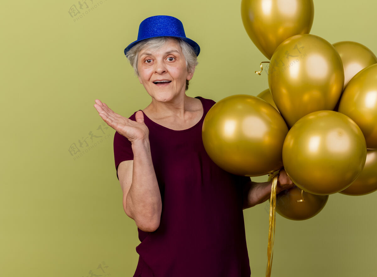 欢乐快乐的老妇人戴着聚会帽站在那里 手里拿着氦气球 手张开放在橄榄绿上绿色聚会橄榄