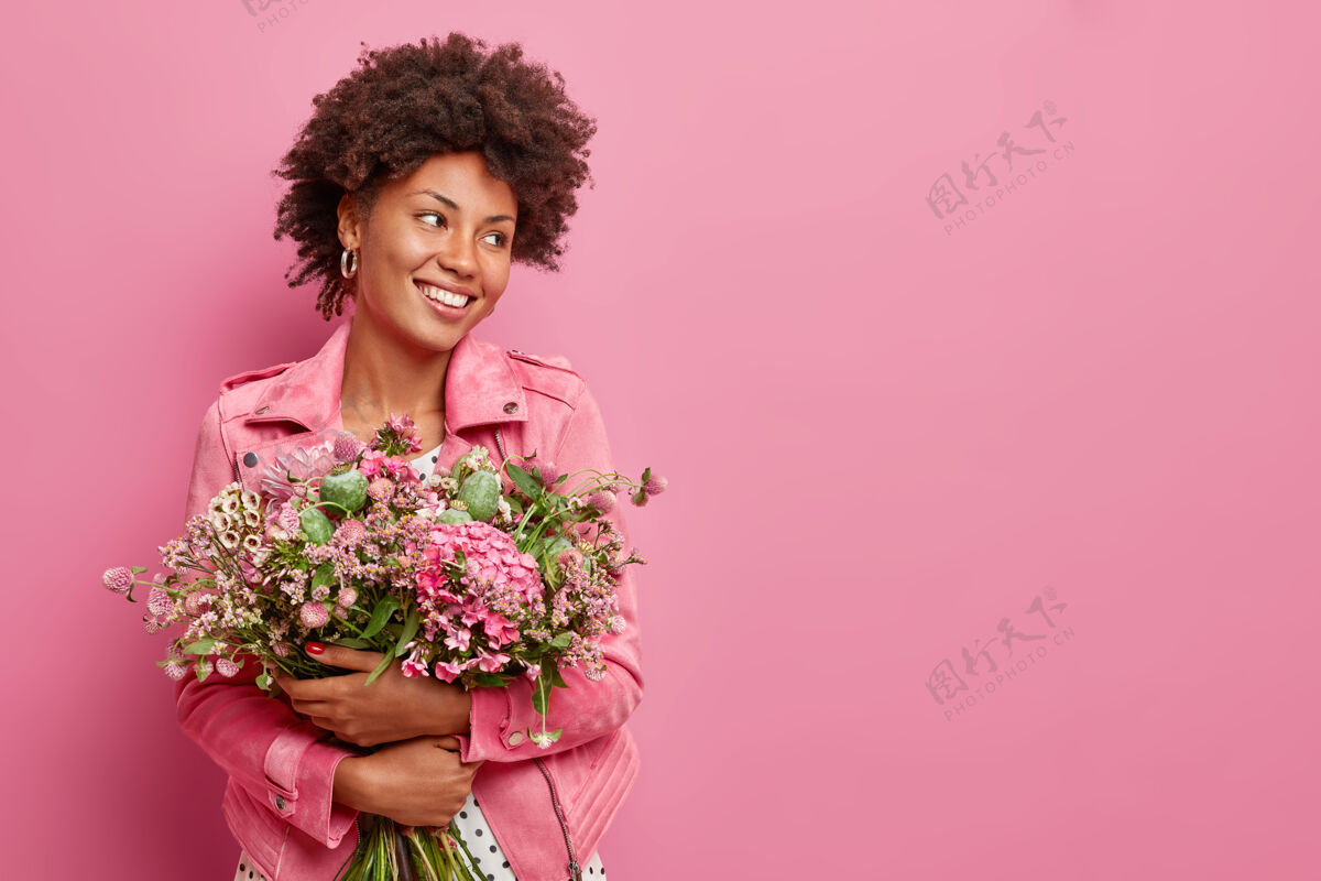 可爱摄影棚拍摄的“快乐女人”手持大束鲜花庆祝春节假期微笑愉快地移开视线对着粉色墙壁摆出姿势并留有复制空间供您推广三月礼物新鲜