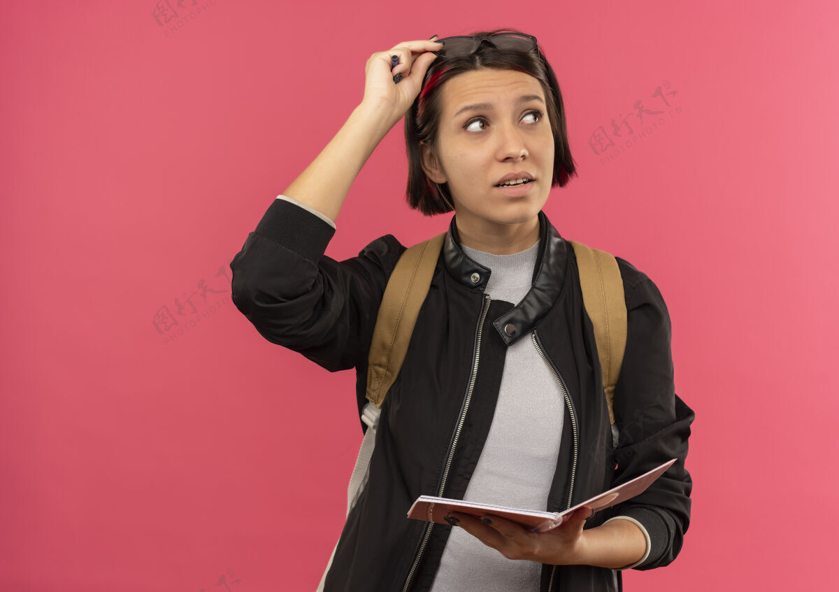 抱体贴的年轻女学生头戴眼镜 背包拿着记事本和眼镜 看着隔离在粉红色墙上的一面衣服思想背
