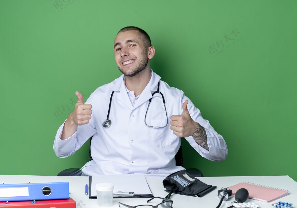人面带微笑的年轻男医生穿着医用长袍 听诊器坐在办公桌旁 工作工具竖起大拇指 隔离在绿色的墙上穿着微笑拇指