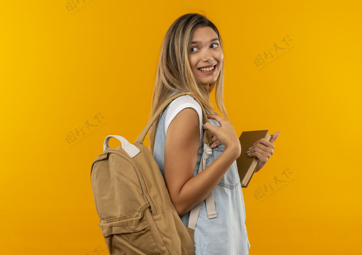 人微笑着的年轻漂亮的学生女孩 背着书包站在侧视图里 手里拿着书 看着她身后孤立的橙色墙壁穿着橙色年轻