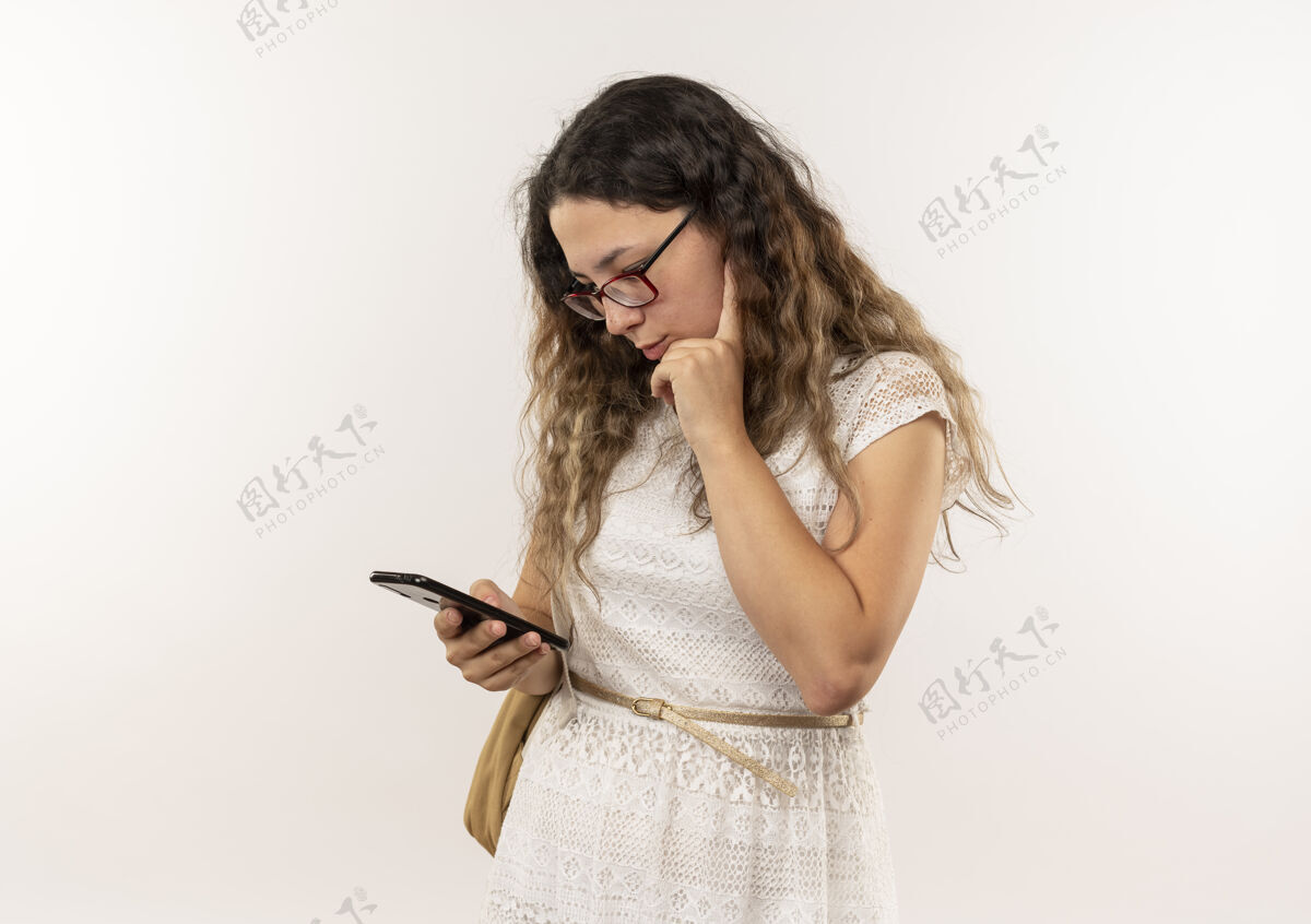 人体贴的年轻漂亮的女学生戴着眼镜 背着书包 用手机把手放在隔离在白墙上的下巴上表情下巴白