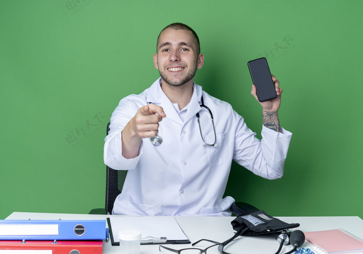 人面带微笑的年轻男医生 穿着医用长袍 手持听诊器 坐在办公桌旁 拿着工作工具 拿着手机 指着隔离在绿色墙上的前面医生穿着表情