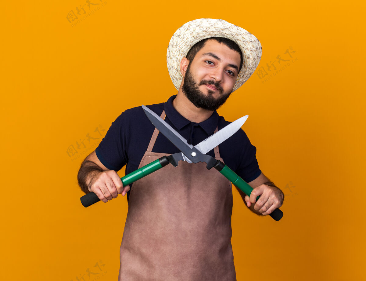 持有高兴的年轻白人男园丁戴着园艺帽手持园艺剪刀隔离在橙色的墙上与复制空间剪刀白种人园艺