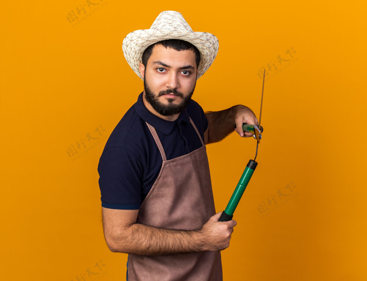 男性严肃的年轻白人男性园丁戴着园艺帽拿着园艺剪刀隔离在橙色的墙上 留着复制空间花园帽子严重