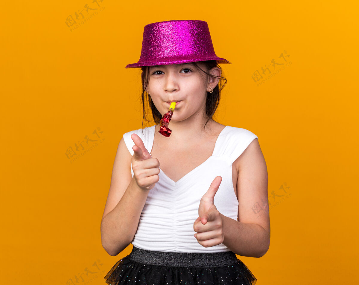 哨子高兴的年轻白人女孩与紫色党帽子吹党哨子 并指出孤立的橙色墙上与复制空间年轻拜托白种人