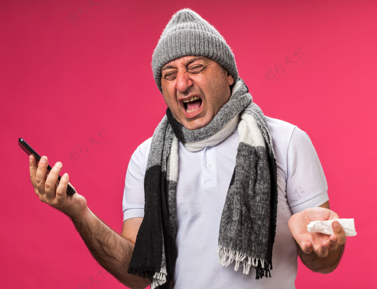 电话恼怒的成年白种人 脖子上围着围巾 戴着冬天的帽子 拿着电话和餐巾 隔离在粉红色的墙上 留着复印空间冬天孤立穿