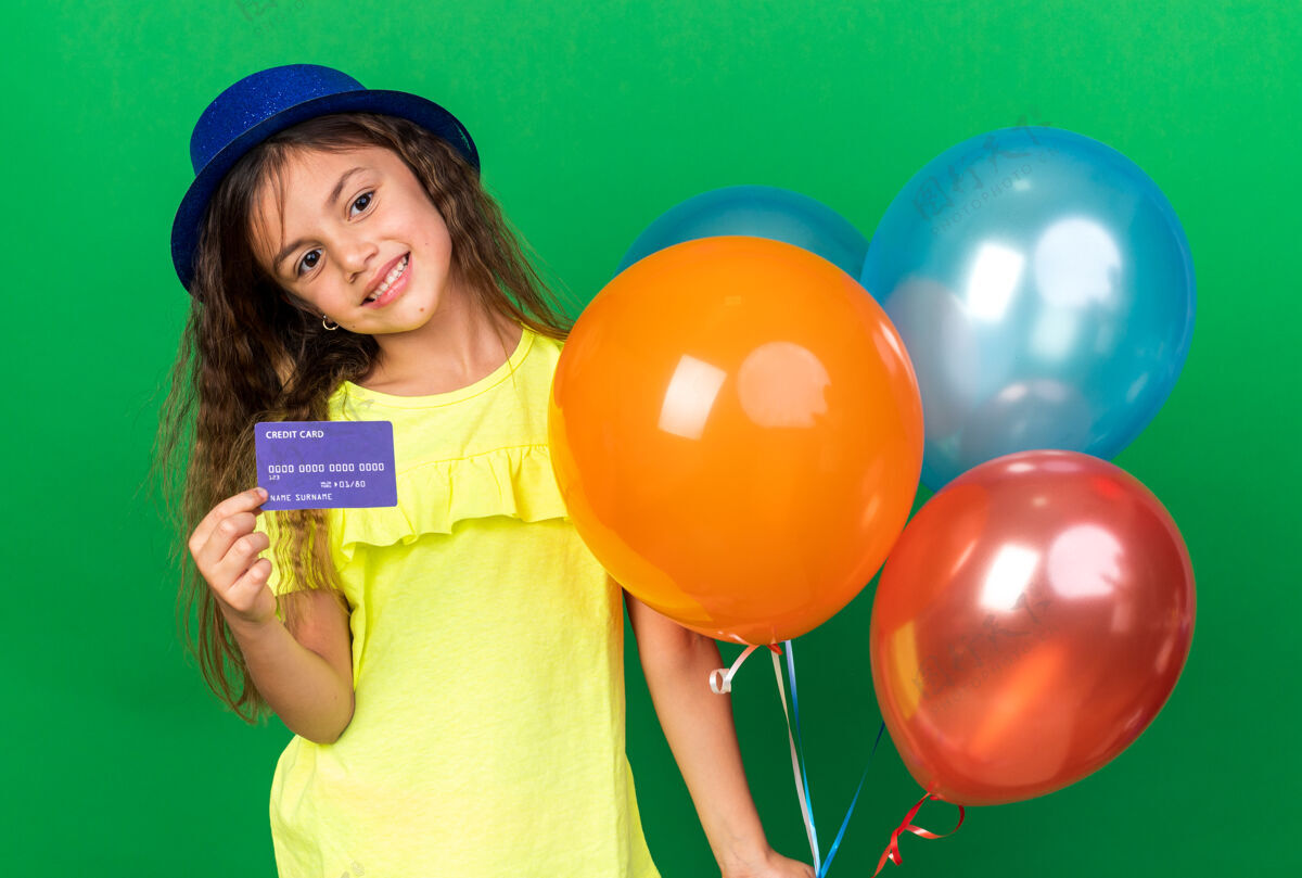 气球可爱的白人小女孩戴着蓝色派对帽 手里拿着氦气球 信用卡被隔离在绿色的墙上 还有复印空间持有生日派对