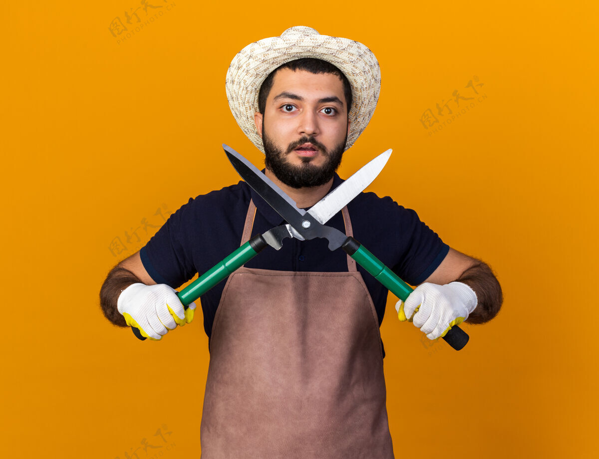 焦虑焦虑的年轻白人男园丁戴着园艺帽和手套拿着园艺剪刀隔离在橙色的墙上 留着复印空间男性剪刀园艺
