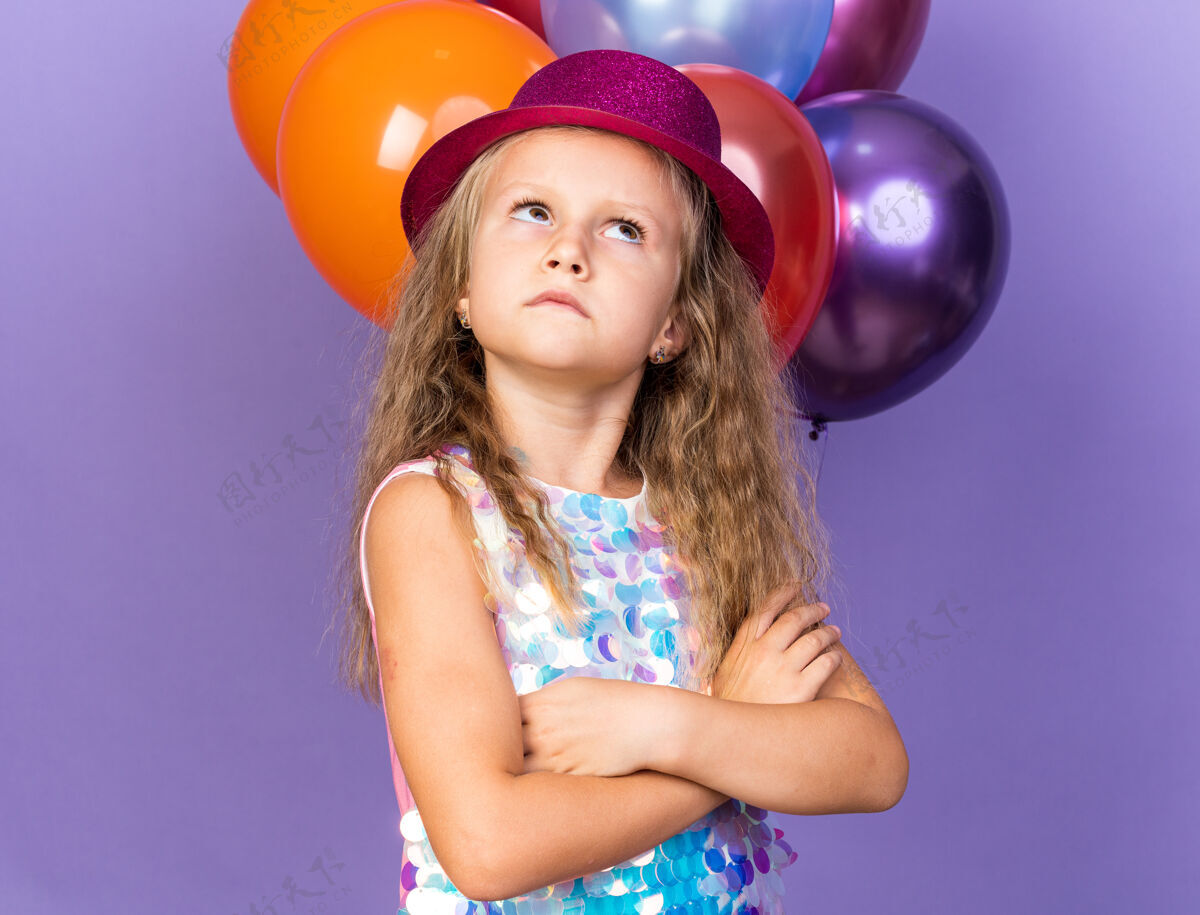 派对恼怒的金发小女孩 戴着紫色的派对帽 双臂交叉站在氦气球前 孤立地看着紫色的墙上有复制空间氦恼火小