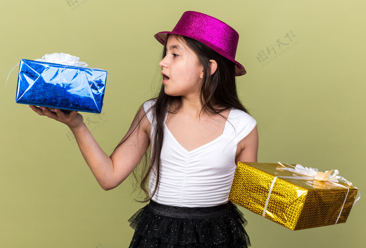 帽子惊讶的年轻白人女孩 戴着紫色派对帽 看着每只手上拿着的礼盒 隔离在橄榄绿的墙上 留着复印空间举行年轻盒子