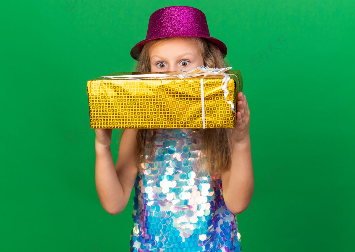 金发惊讶的金发小女孩 戴着紫色派对帽 拿着礼品盒 隔离在绿色的墙上 还有复制空间小女孩派对