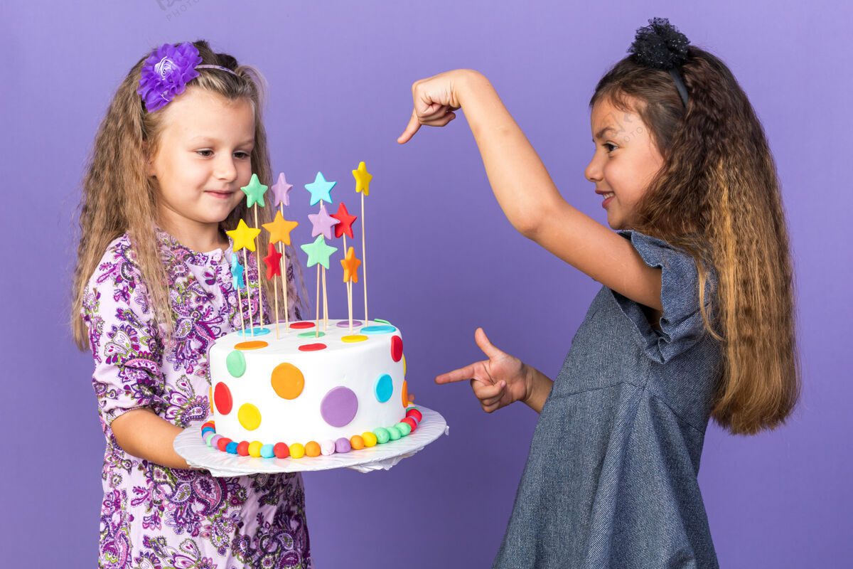 抱着笑眯眯的白人小女孩指着拿着生日蛋糕的金发小女孩 这个蛋糕被隔离在紫色的墙上 还有一个复制空间孤立的白种人指着