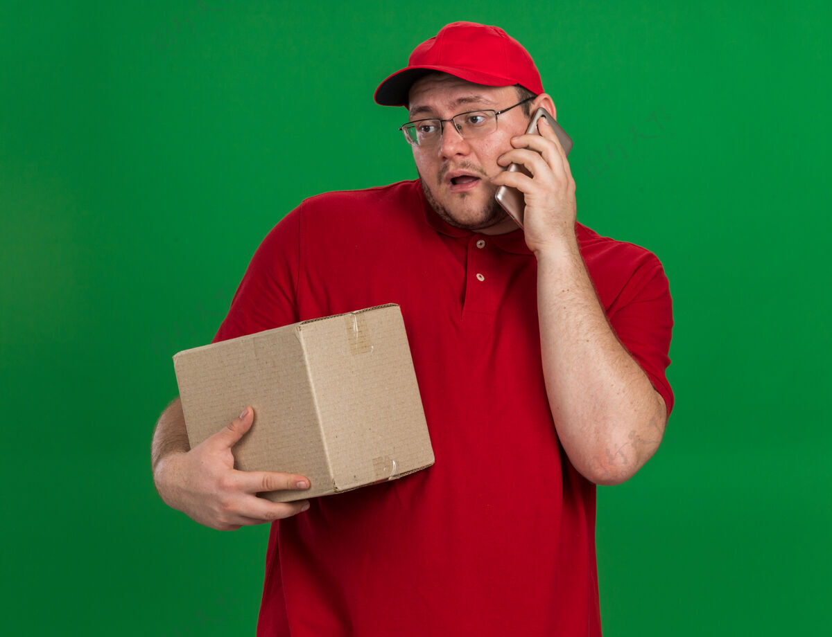 超重失望超重的年轻送货员 戴着眼镜 拿着纸板箱 讲着电话 隔离在绿色的墙上 留着复印空间手持年轻光学