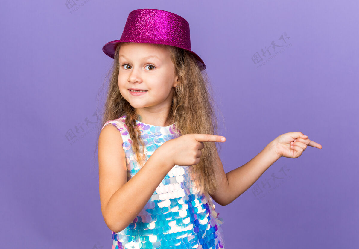 女孩令人印象深刻的小金发女孩与紫罗兰党的帽子指向一边孤立的紫色墙上复制空间生日印象壁板