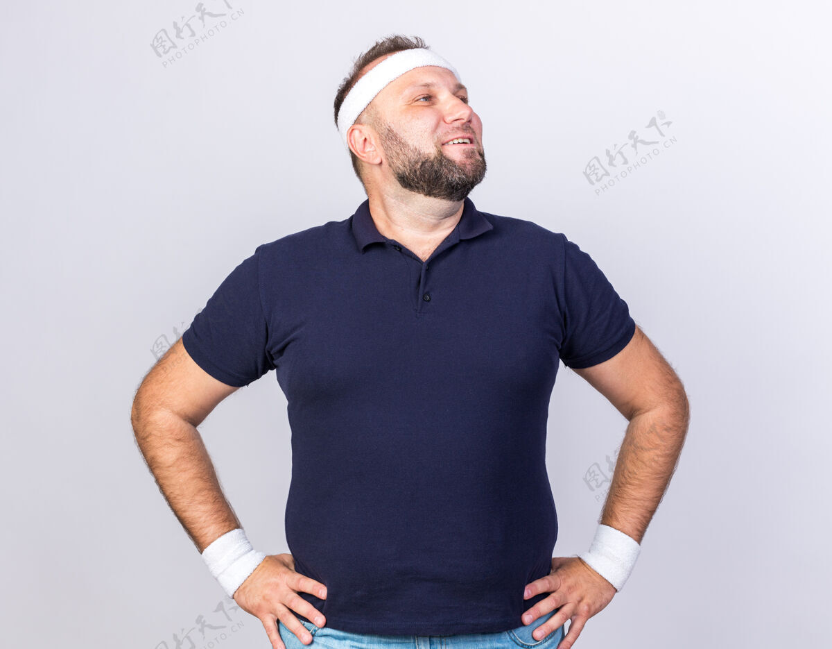 穿面带微笑的成年斯拉夫运动型男子戴着头带和腕带 双手放在腰上 看着隔离在白色墙壁上的一面 留着复制空间斯拉夫运动手