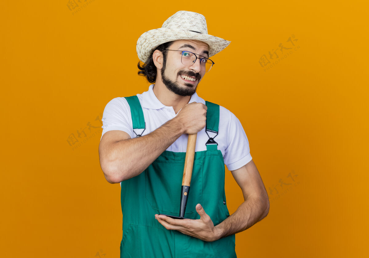 表情年轻的留着胡须的园丁 穿着连体衣 戴着帽子 拿着小耙子 站在橙色的墙上 兴高采烈地微笑着看着前面胡须连身衣帽子