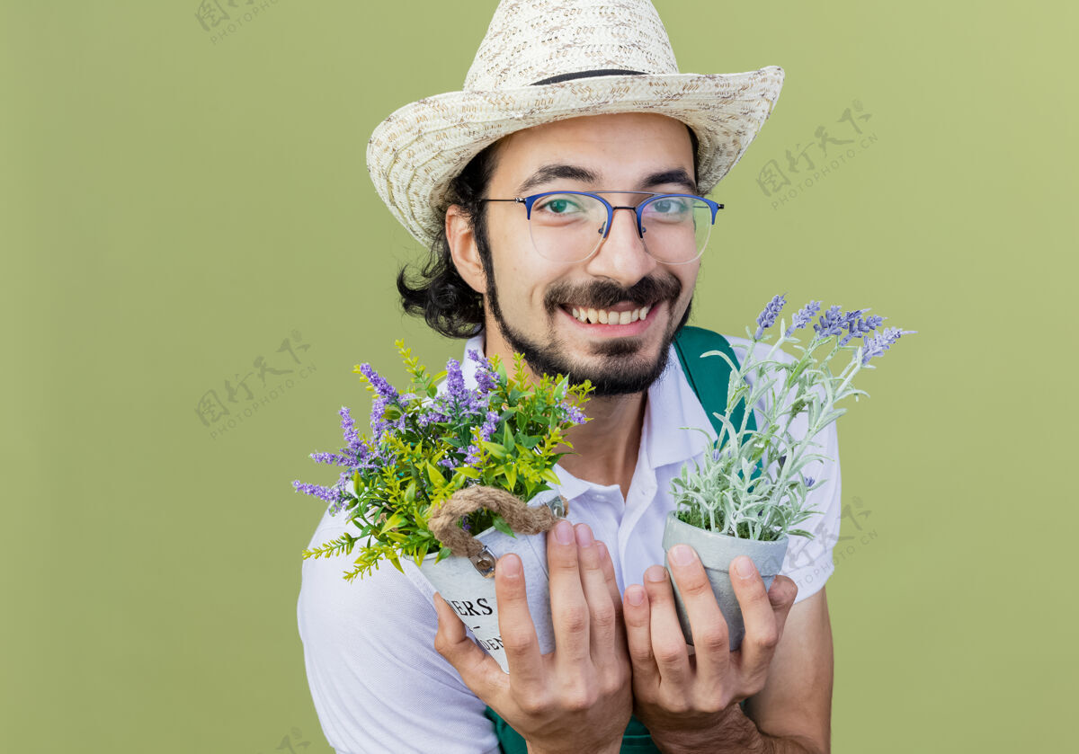 脸年轻的留着胡须的园丁 穿着连身衣 戴着帽子 手里拿着盆栽植物 站在浅绿色的墙上 兴高采烈地微笑着看着前方锅人花园