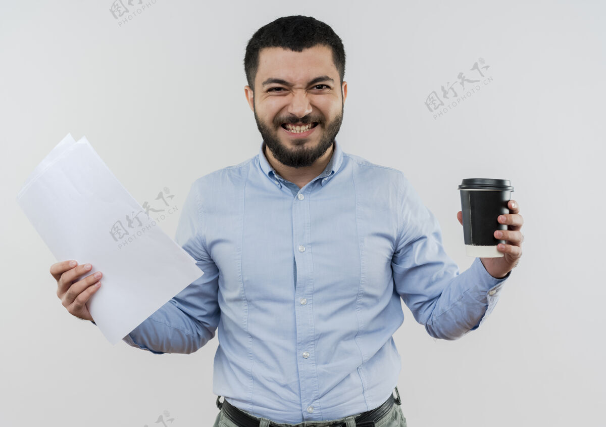 人身穿蓝衬衫 留着胡子的年轻人 手里拿着咖啡杯 站在白墙上 高兴而兴奋咖啡文件拿着