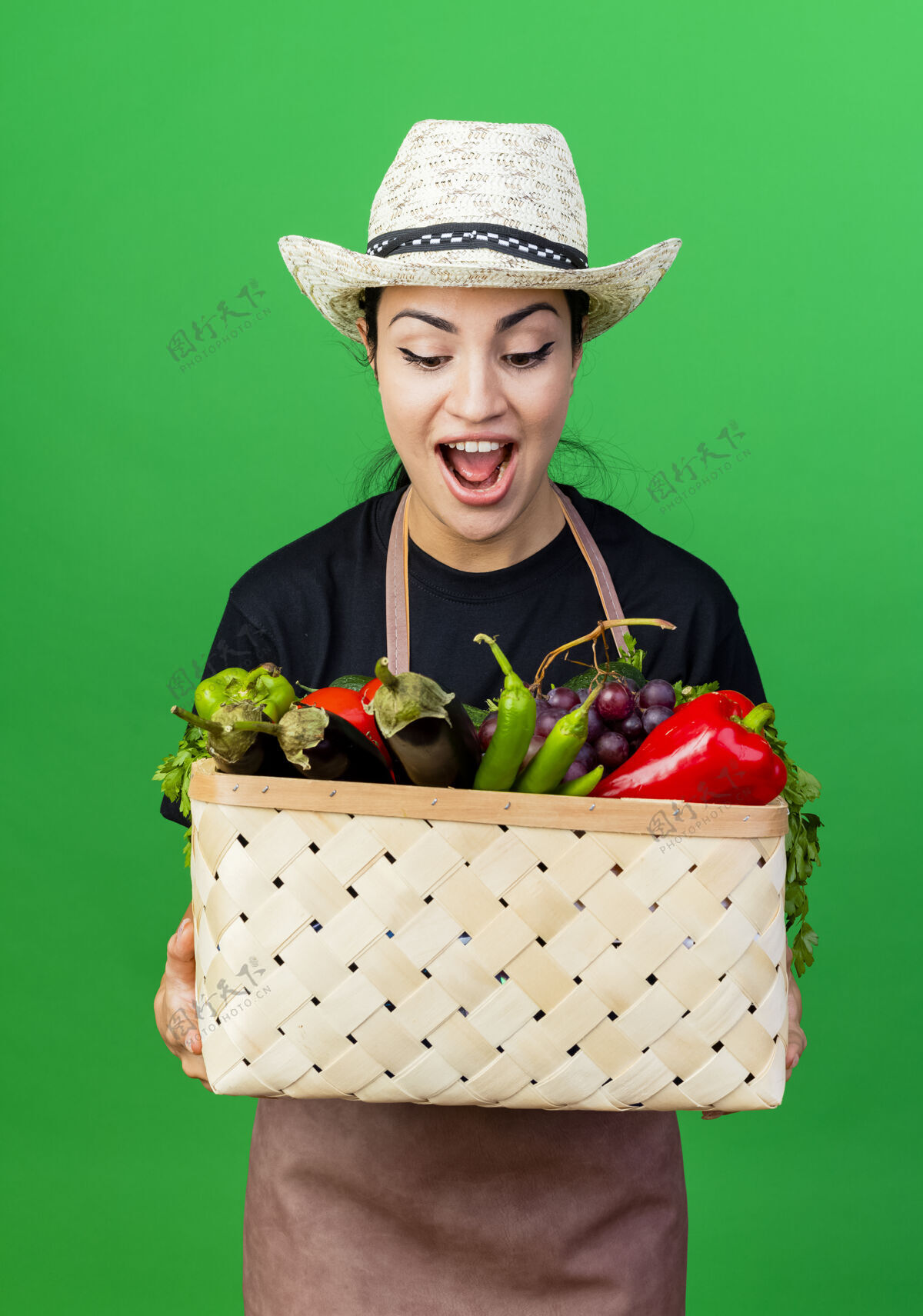 围裙年轻漂亮的女园丁 围着围裙 戴着帽子 手里拿着装满蔬菜的篮子 微笑着看着它 站在绿色的墙上 惊讶地笑着女人脸微笑