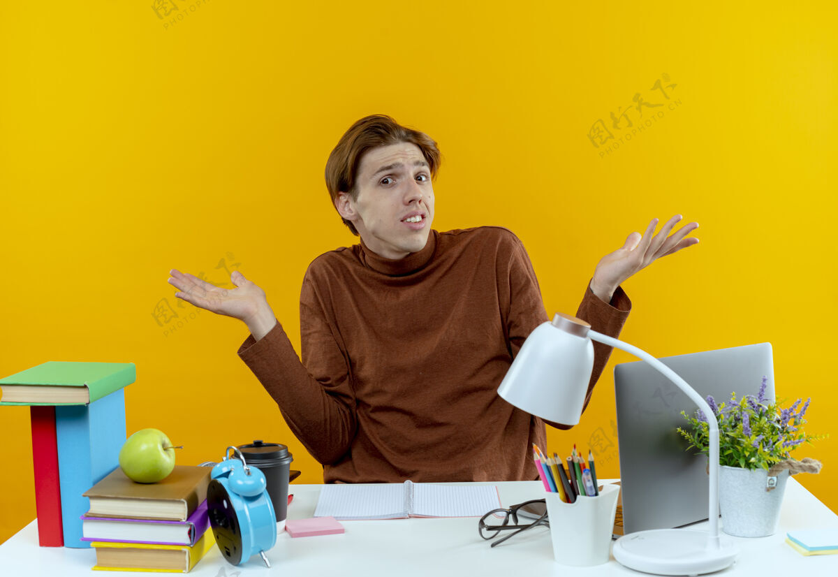 学生困惑的年轻学生男孩坐在书桌旁 手里拿着学习用具摊开双手困惑书桌黄色