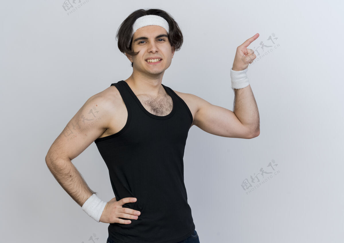 脸年轻的运动型男子穿着运动服 头戴头巾 面带微笑 自信地用食指指着站在白墙上的一边自信衣服运动