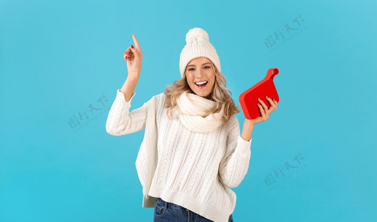 女人时尚的金发微笑的美丽年轻女子手持无线扬声器听音乐穿着白色毛衣和针织帽子摆在蓝色衣服女士舞蹈