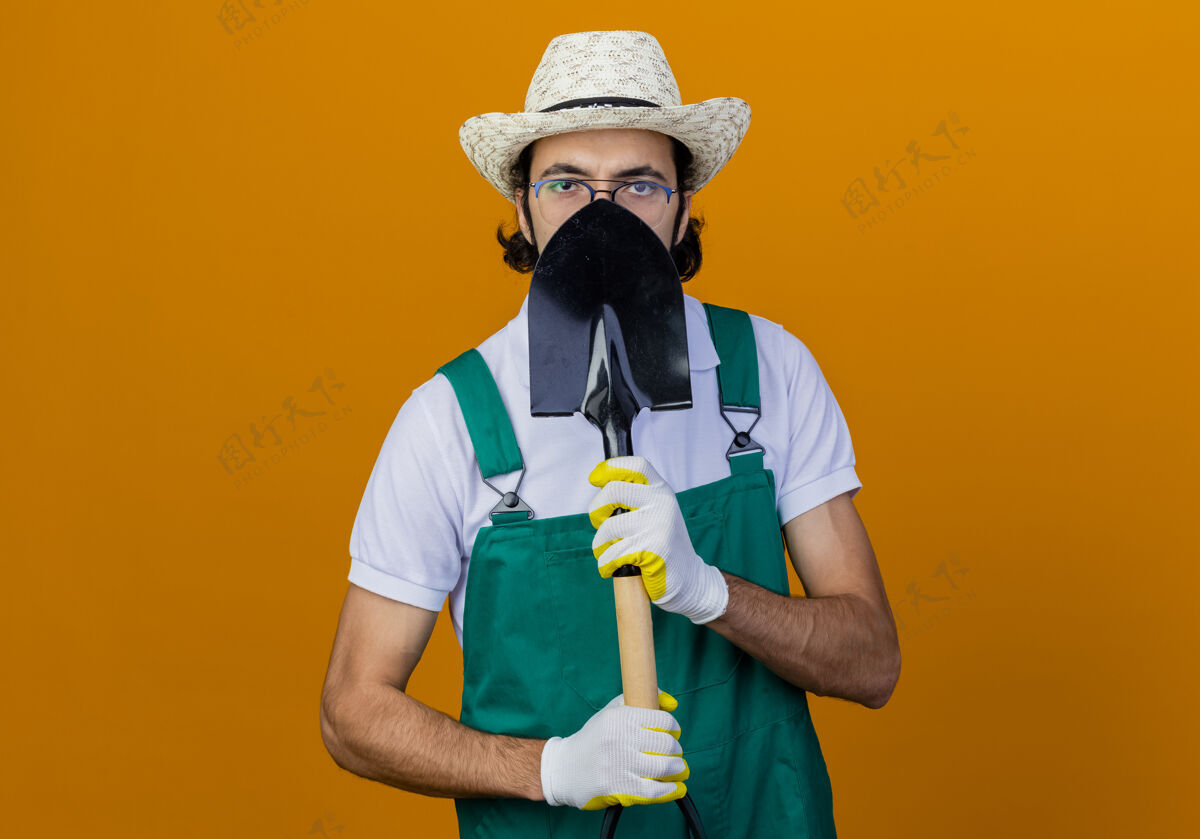 人年轻的留着胡子的园丁 穿着连体衣 戴着帽子 手里拿着铲子 站在橙色的墙上遮住了脸脸花园藏