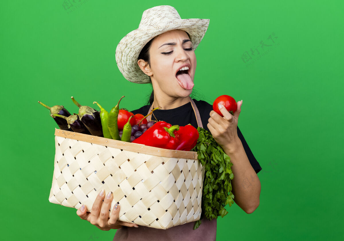 女人年轻漂亮的女园丁围着围裙 戴着帽子 手里拿着装满蔬菜的篮子 站在绿色的墙上 带着厌恶的表情看着西红柿站着西红柿表情