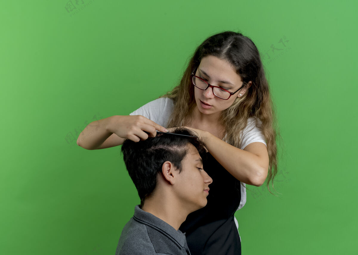 梳子年轻的专业美发师 围裙里的女人 站在绿墙上梳男顾客的头发姿势年轻人人