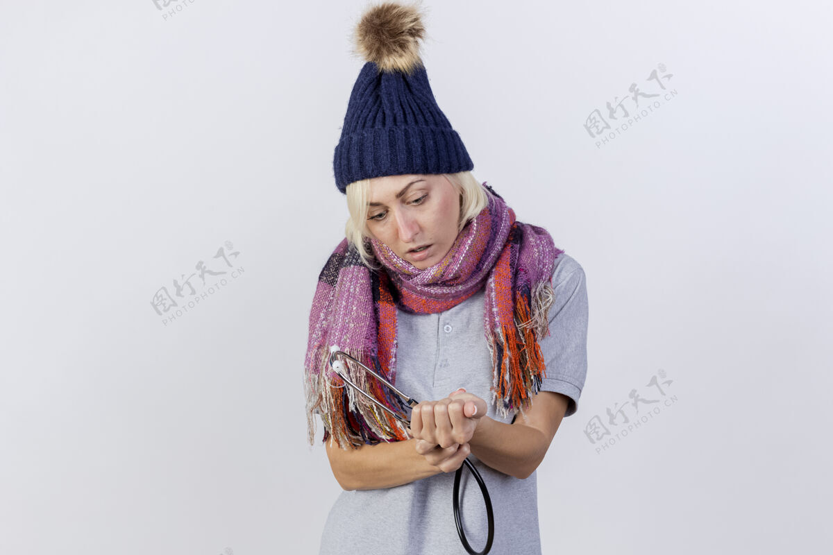 人令人印象深刻的年轻金发病妇女戴着冬季帽子和围巾手持听诊器 看着隔离在白墙上的手人疾病表情