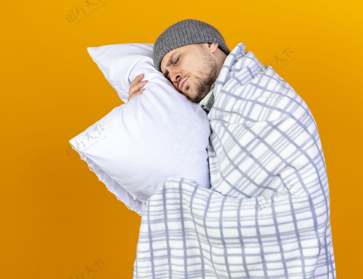 帽子昏昏欲睡的年轻金发病夫戴着冬天的帽子 围巾裹着格子布 把头靠在隔离在橙色墙上的枕头上金发脸姿势