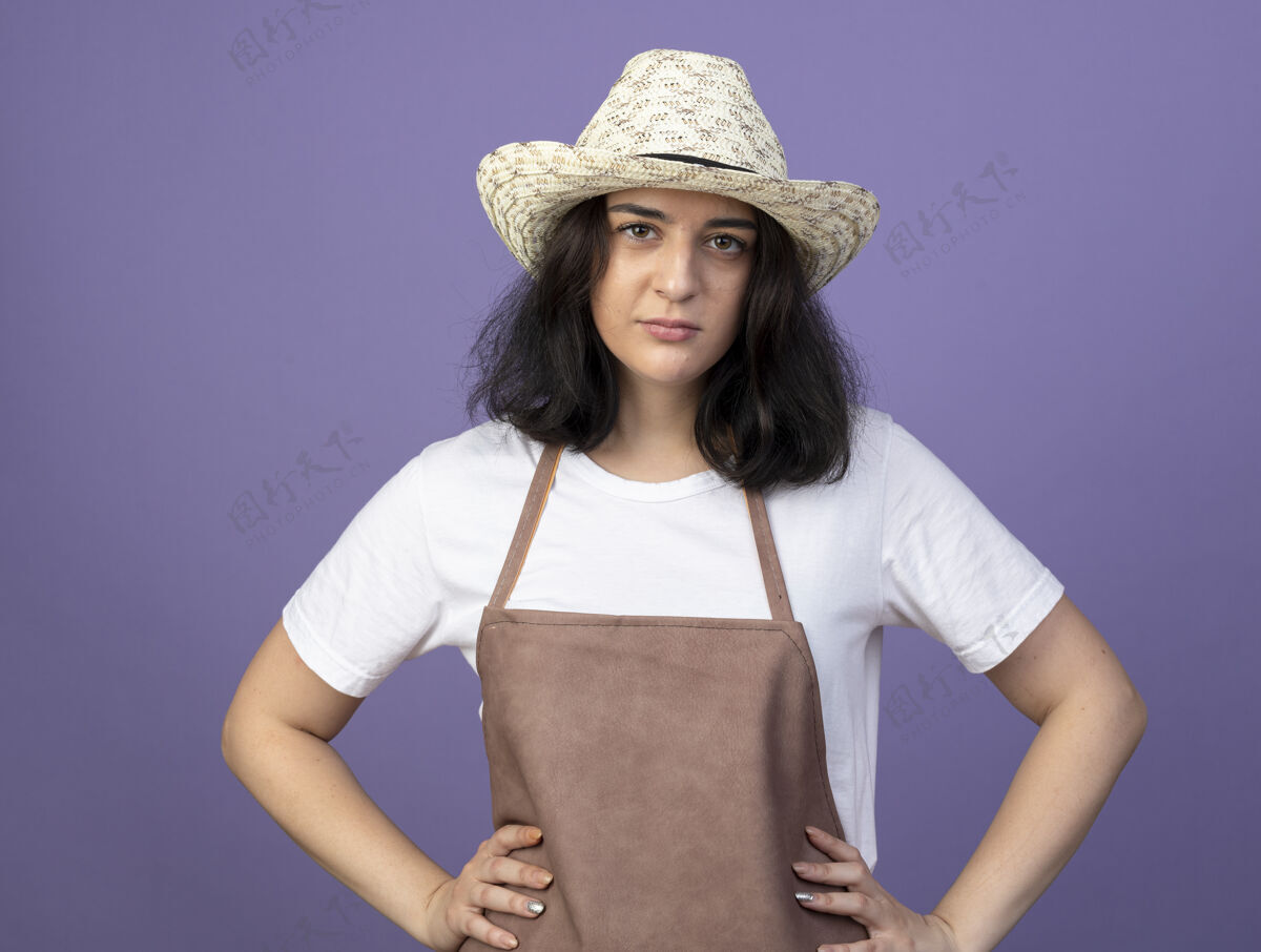 脸自信的年轻黑发女园丁穿着制服 戴着园艺帽 双手放在腰上 看着紫色墙上孤立的前方自信年轻人腰
