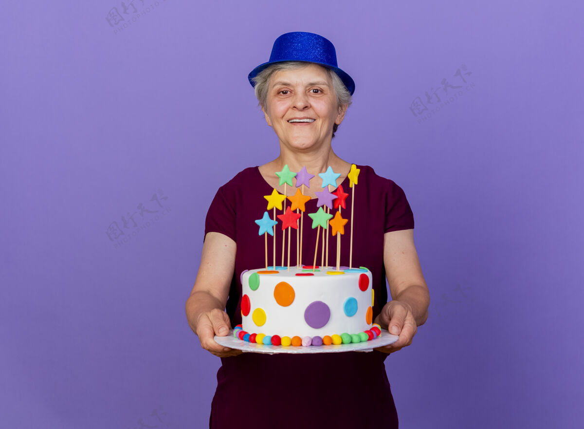 聚会戴着派对帽的快乐的老妇人在紫色的墙上举着生日蛋糕女人脸生日
