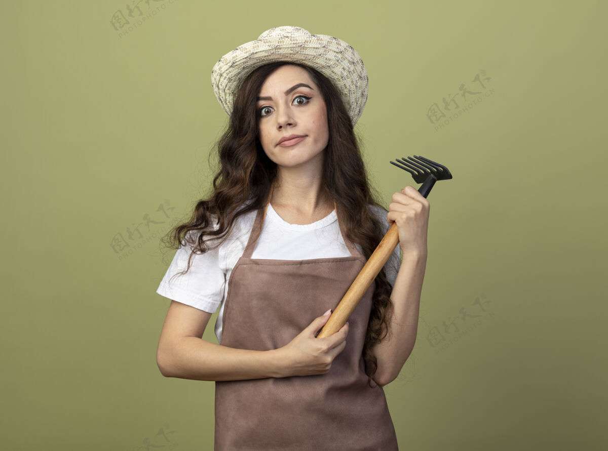 帽子年轻的女园丁穿着制服 戴着园艺帽 拿着耙子 望着橄榄绿墙上孤立的前方脸印象耙子